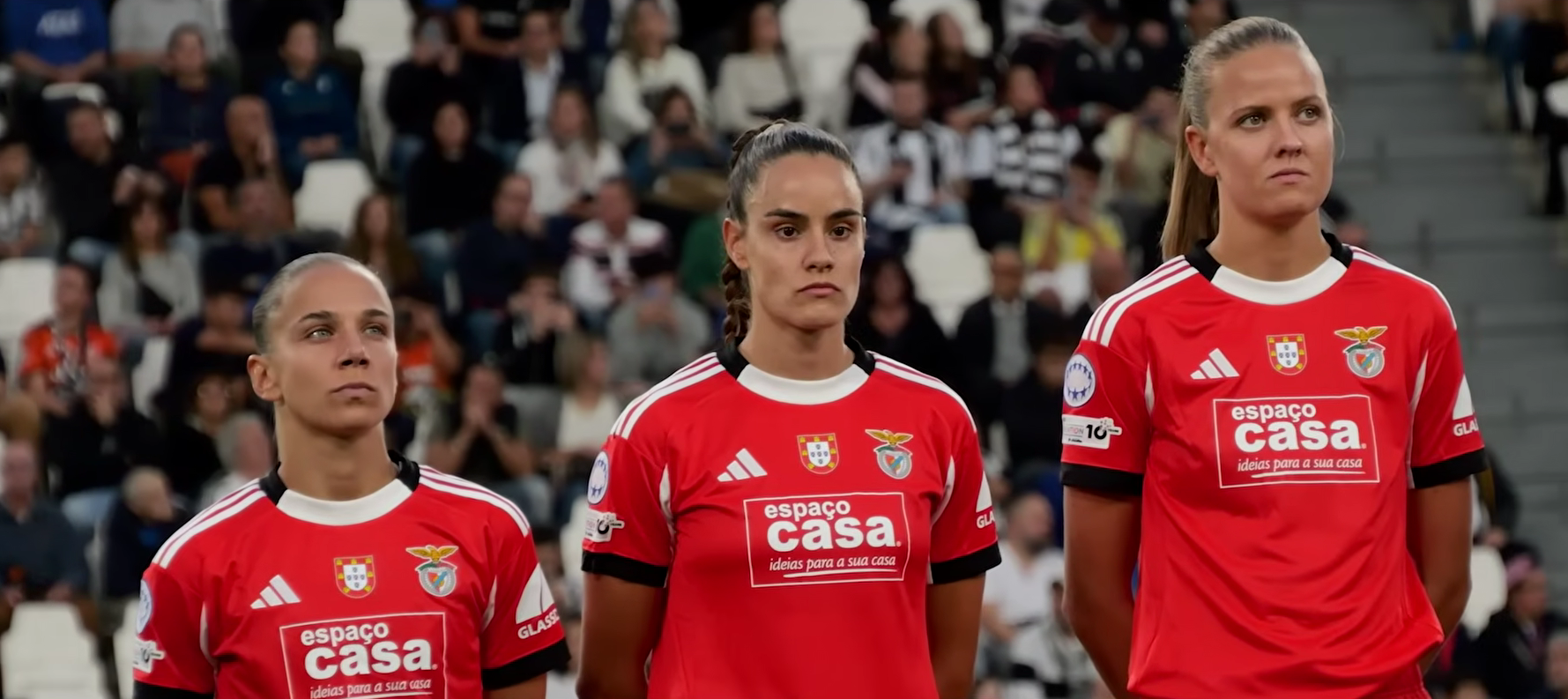 Három Benfica-játékos piros mezben áll a stadion lelátója előtt.